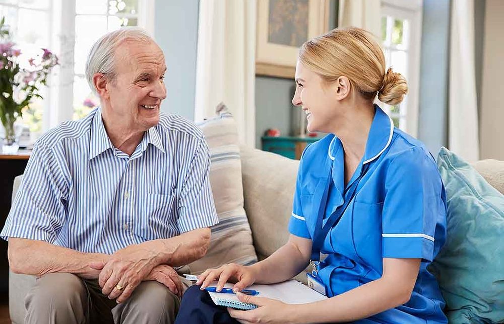 A nurse in blue scrubs smiling and talking with an older man during a friendly in-home visit